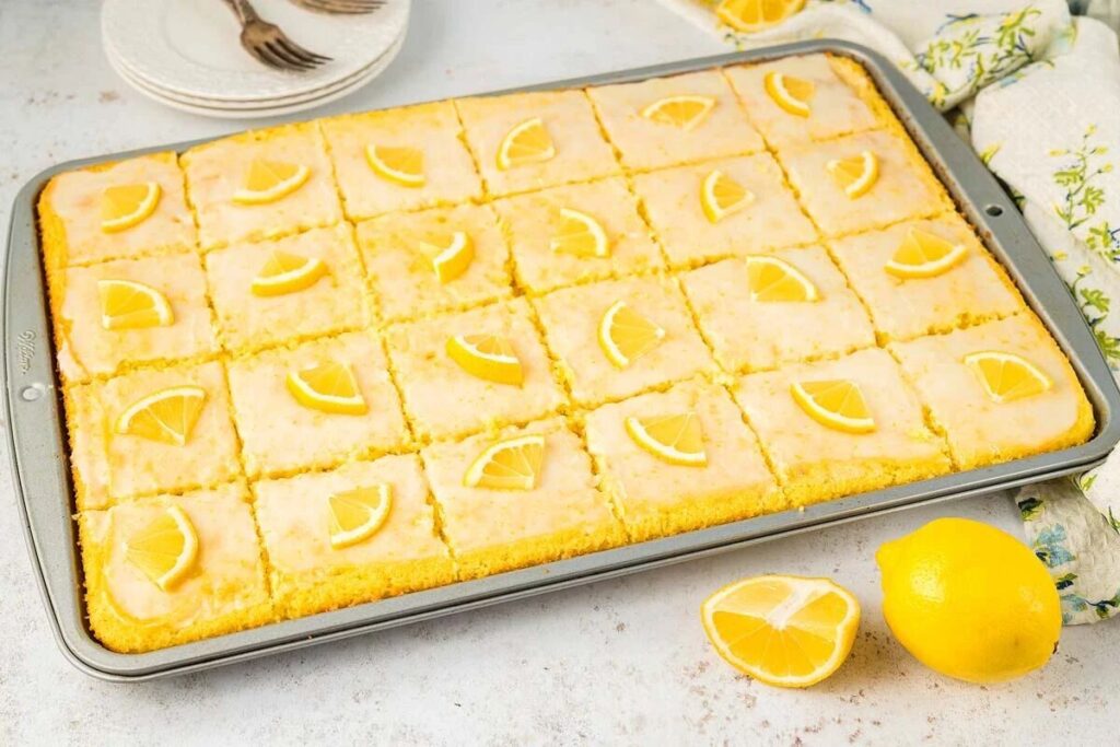 A tray of lemon sheet cake features slices garnished with lemon wedges, surrounded by fresh lemons and serving utensils.