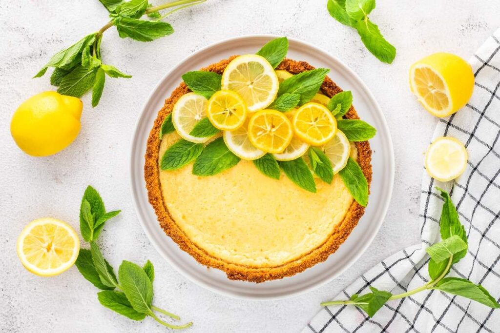 A lemon tart with mint and lemon slices rests among fresh lemons and mint leaves.