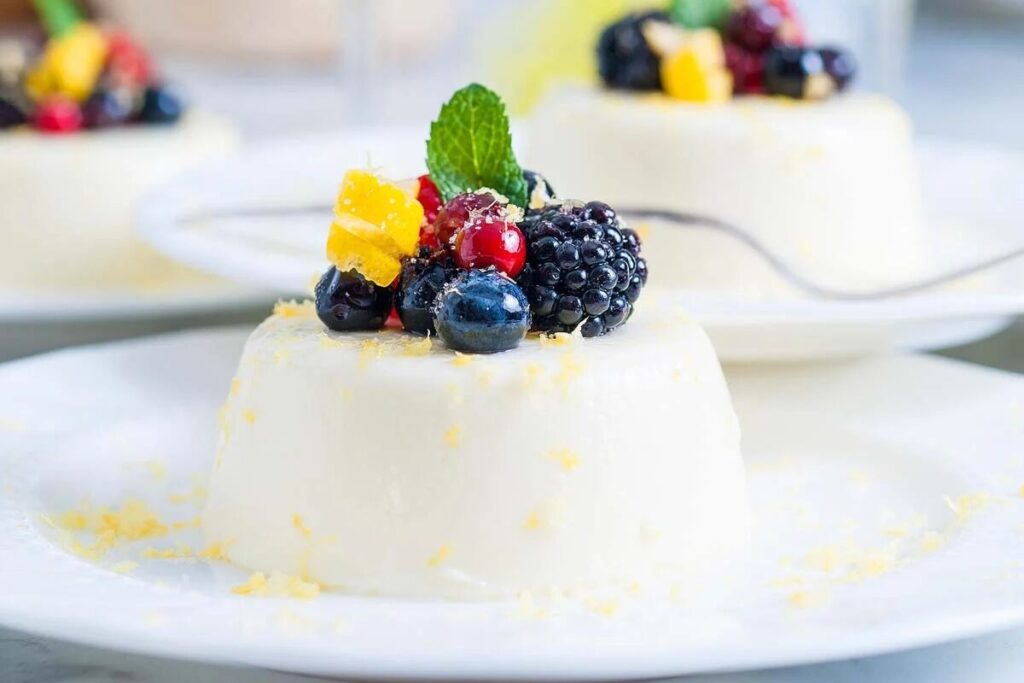 A creamy panna cotta topped with vibrant berries, a sprig of mint, and lemon zest on a white plate.