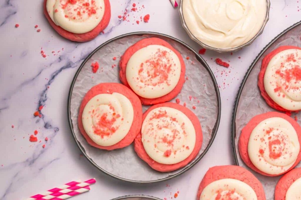 Overhead shot of several pink frosted sugar cookies on metal trays, along with a bowl of frosting and two pink and white striped straws, all arranged on a white marble surface. The cookies have a thick swirl of white frosting in the center, and red sprinkles.
