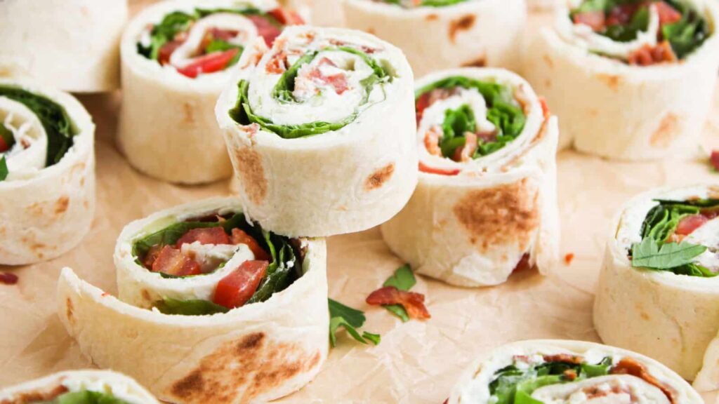 Close-up of tortilla pinwheels filled with leafy greens, tomato, and possibly cream cheese, arranged on parchment paper.