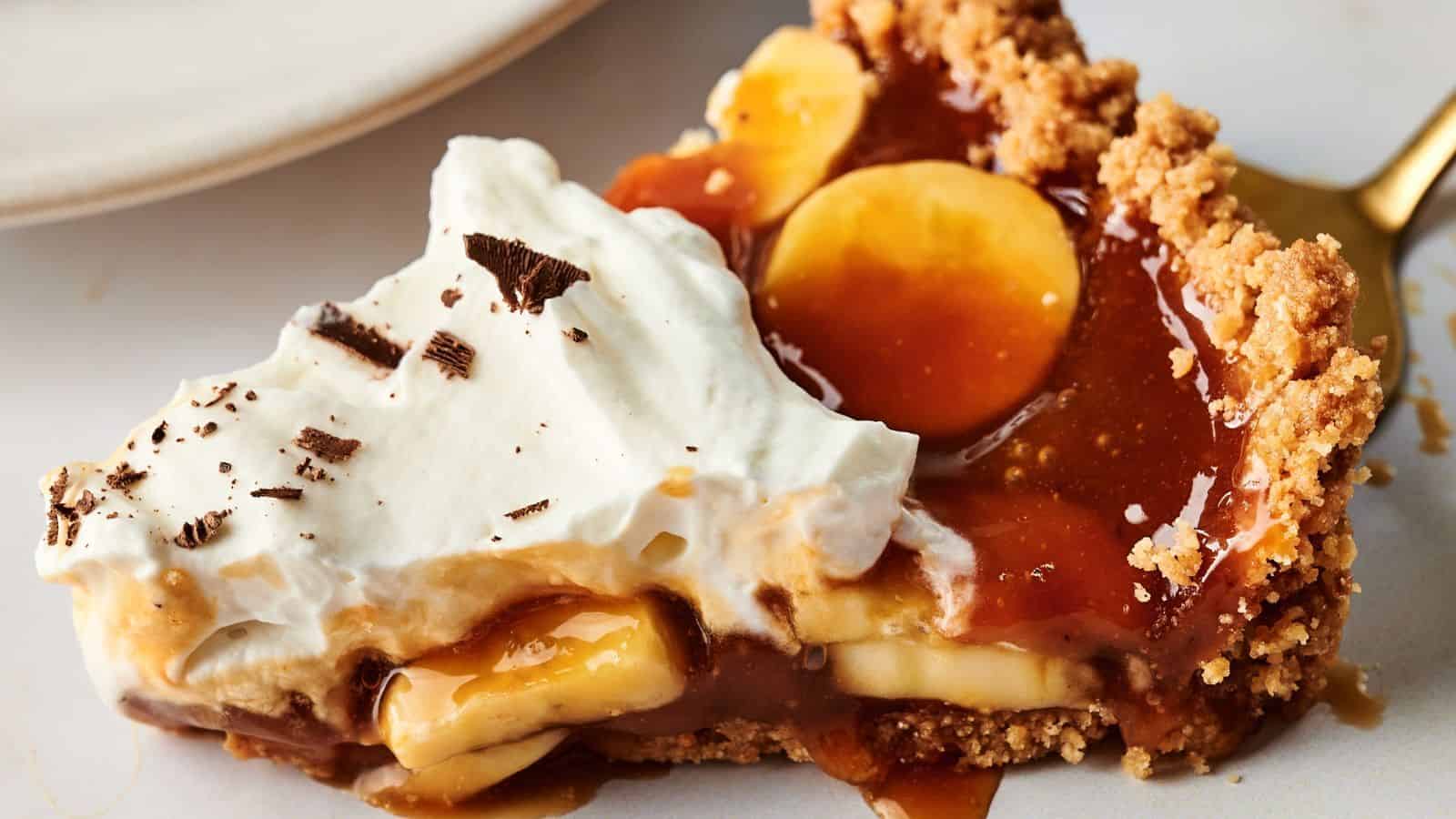 A slice of banoffee pie with layers of banana, caramel, whipped cream, and a crumbly biscuit crust on a white plate.