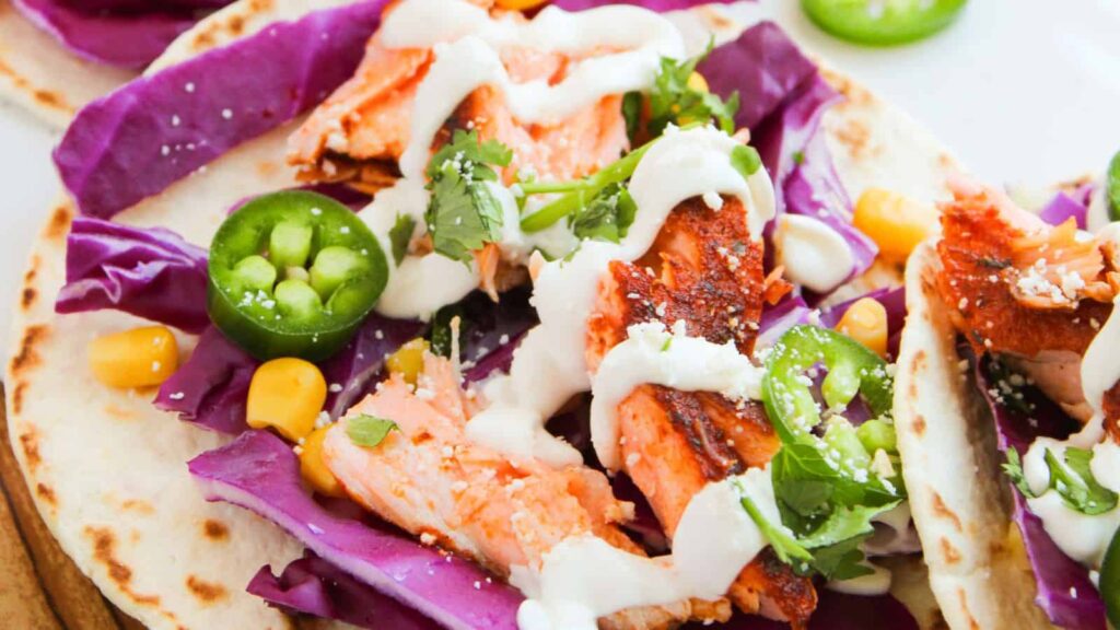 Close-up of tacos filled with grilled salmon, red cabbage, corn, cilantro, sliced jalapeño, and drizzled with white sauce on soft tortillas.