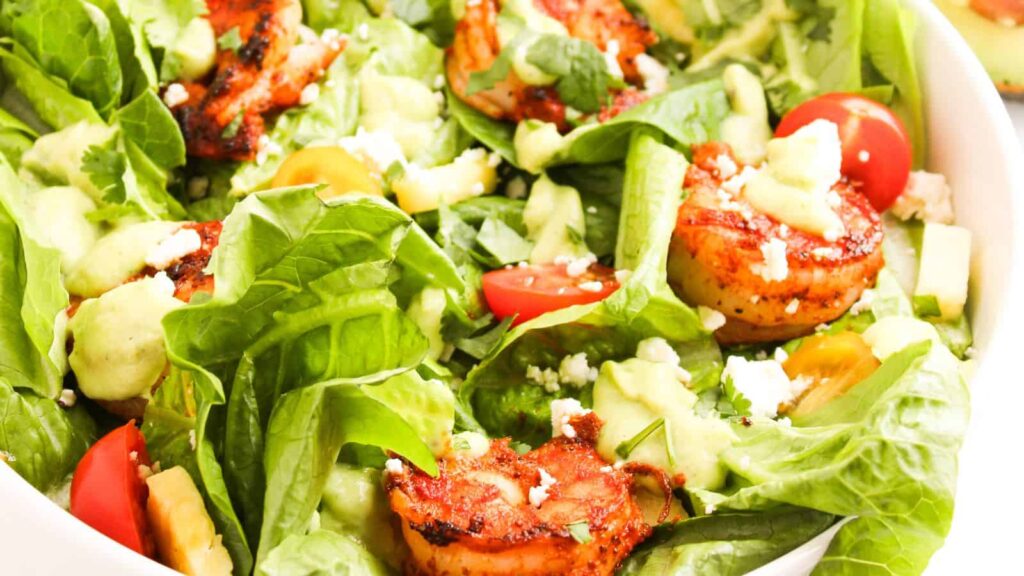 A fresh salad features leafy greens, grilled shrimp, cherry tomatoes, avocado slices, and crumbled cheese, all tossed with a light dressing.