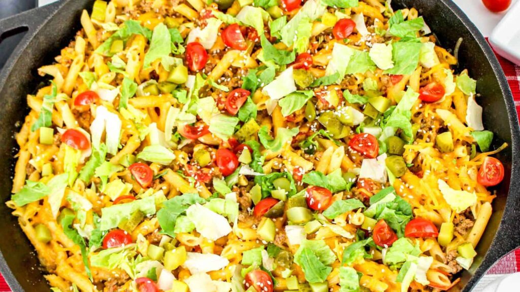 A skillet filled with cooked pasta is topped with chopped lettuce, diced tomatoes, melted cheese, and various other vegetables.