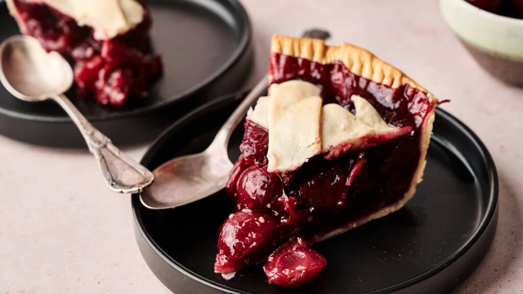 A slice of cherry pie with a golden lattice crust sits on a black plate with a silver spoon beside it.