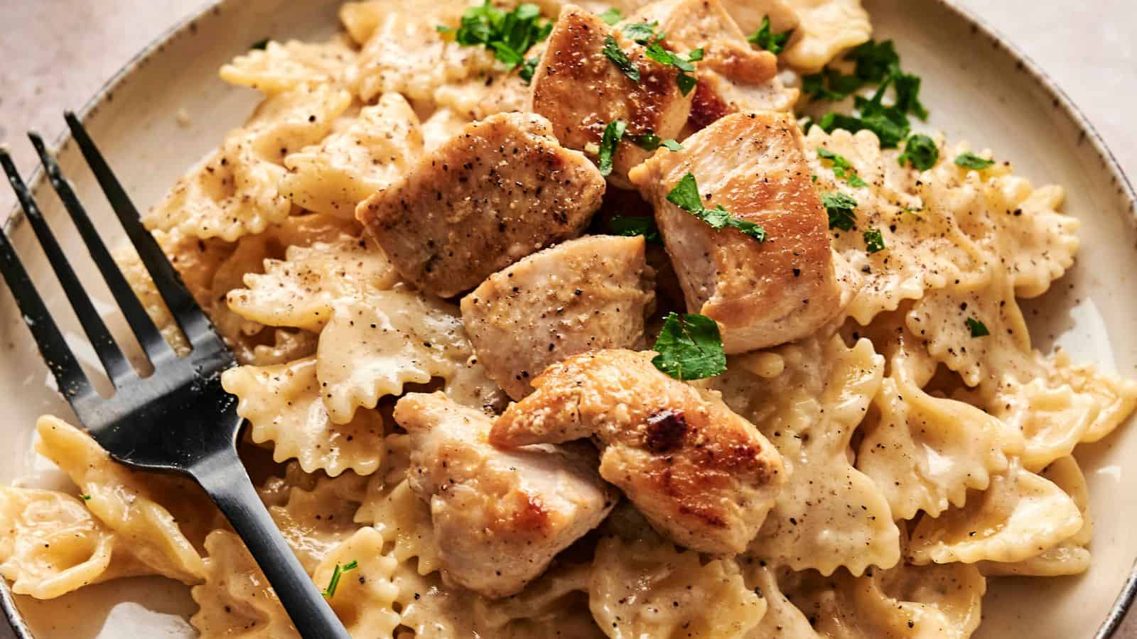 A plate of bowtie pasta with creamy sauce topped with cooked chicken pieces and garnished with parsley, with a black fork on the side.