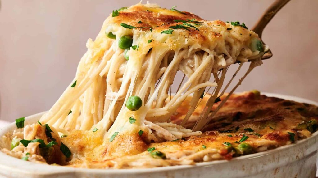 A fork lifts a cheesy serving of baked pasta with peas and shredded chicken from a casserole dish, topped with melted cheese and herbs.