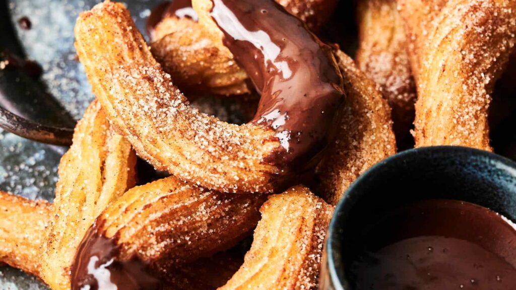 A close-up of churros coated in cinnamon sugar, some dipped in chocolate sauce, with a bowl of chocolate sauce nearby.