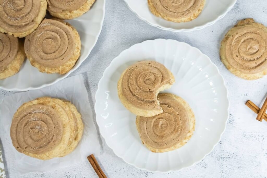 Overhead view of several churro cookies on white plates, one cookie partially eaten, with cinnamon sticks scattered nearby.
