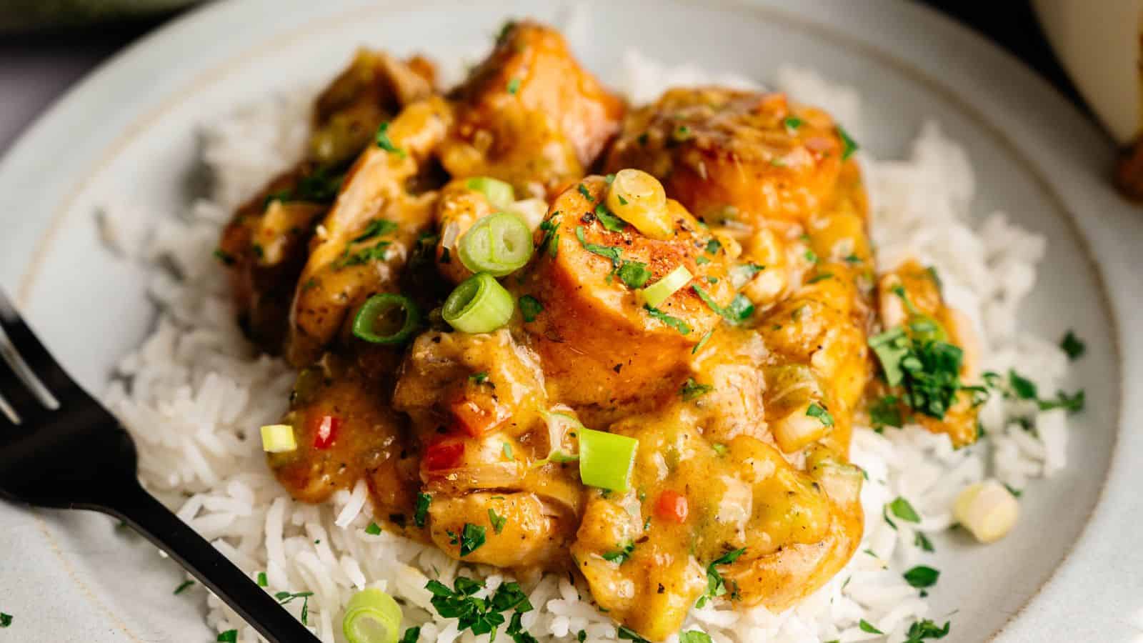 A plate of white rice topped with chunks of chicken in a yellow curry sauce, garnished with chopped green onions and herbs. A black fork is placed on the side.