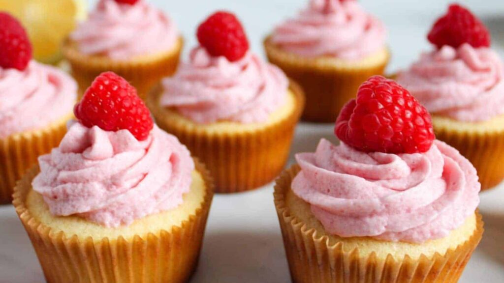A close-up of nine vanilla cupcakes topped with pink frosting and fresh raspberries on a white surface.