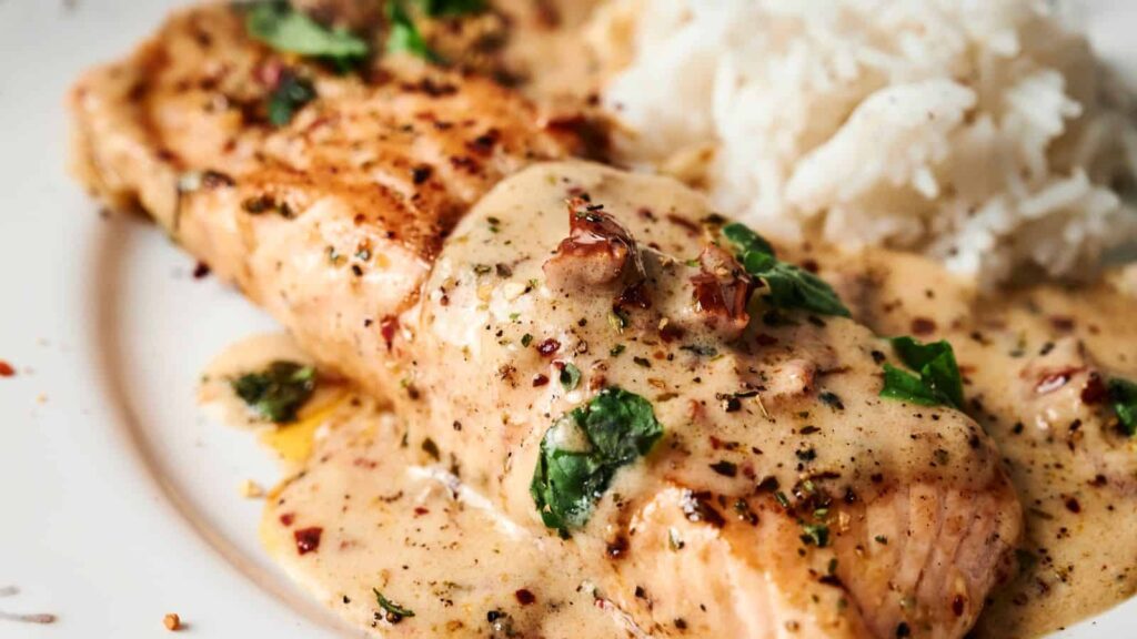 Salmon fillet topped with creamy herb sauce, garnished with herbs, served alongside a portion of white rice on a plate.