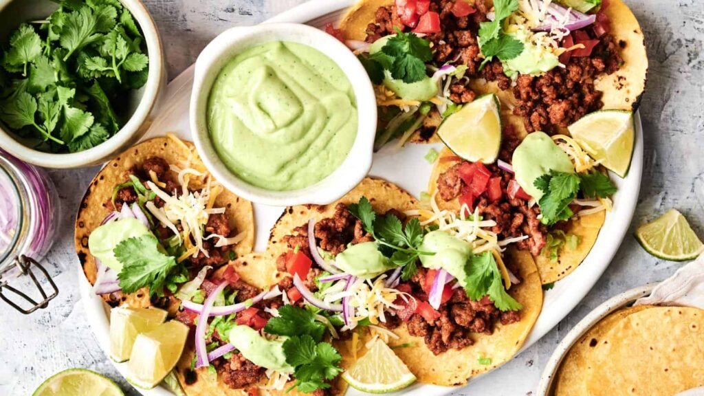 A plate of ground meat tacos topped with shredded cheese, diced tomatoes, red onion, cilantro, and avocado sauce, served with lime wedges and a bowl of green sauce.