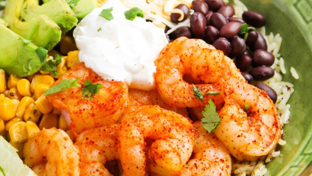 A bowl with seasoned shrimp, black beans, corn, avocado, sour cream, rice, and garnished with cilantro.