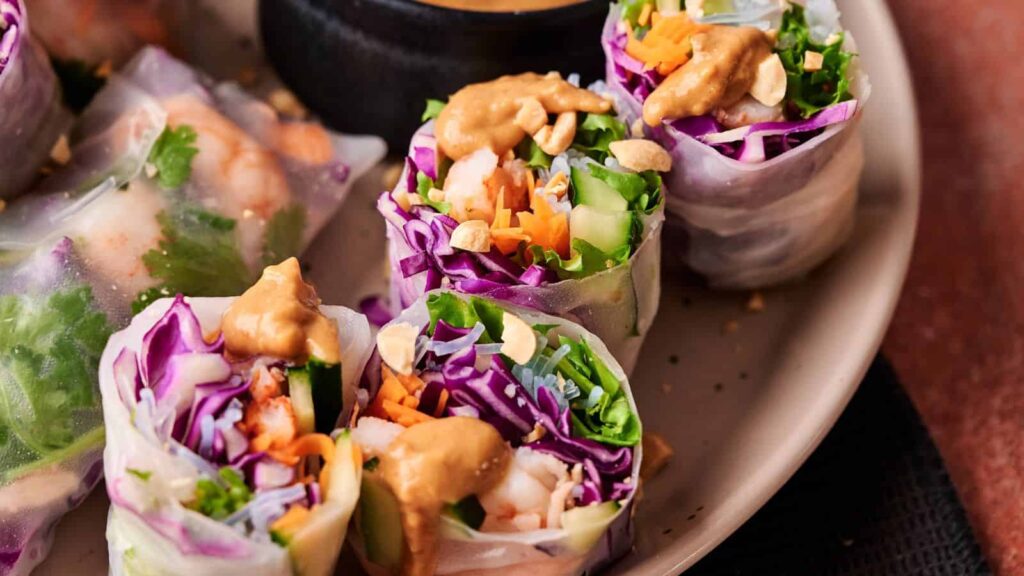 Fresh spring rolls filled with shrimp, lettuce, purple cabbage, carrots, and cucumber, topped with peanut sauce and crushed peanuts, served on a plate with a dipping sauce.