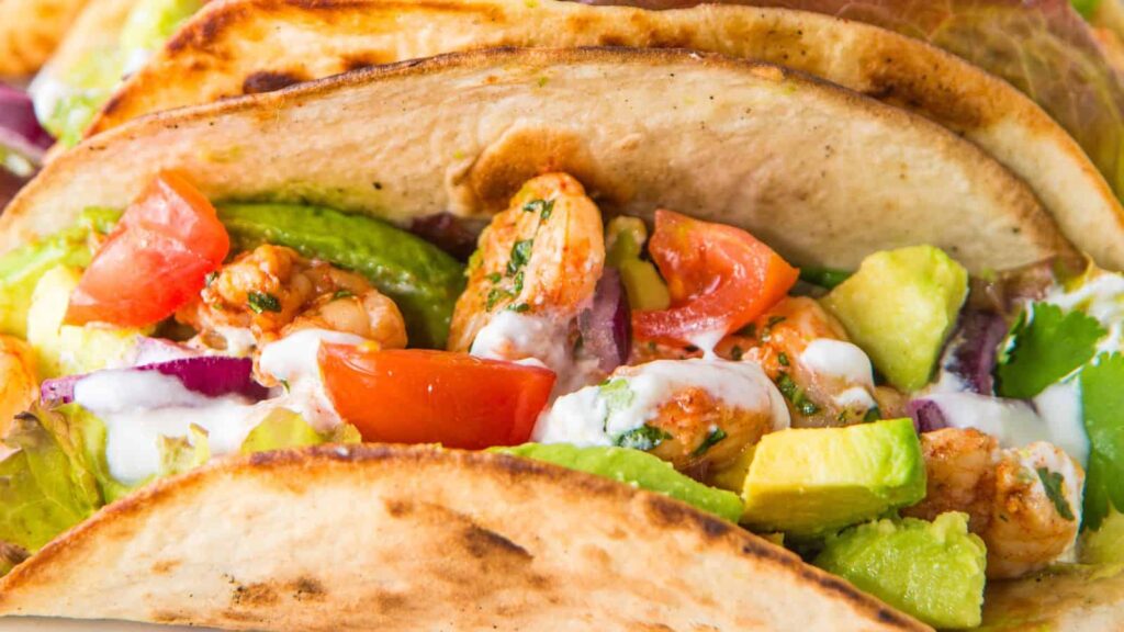 Close-up of a soft-shell taco filled with shrimp, diced tomatoes, avocado, red onion, and drizzled with a white sauce.