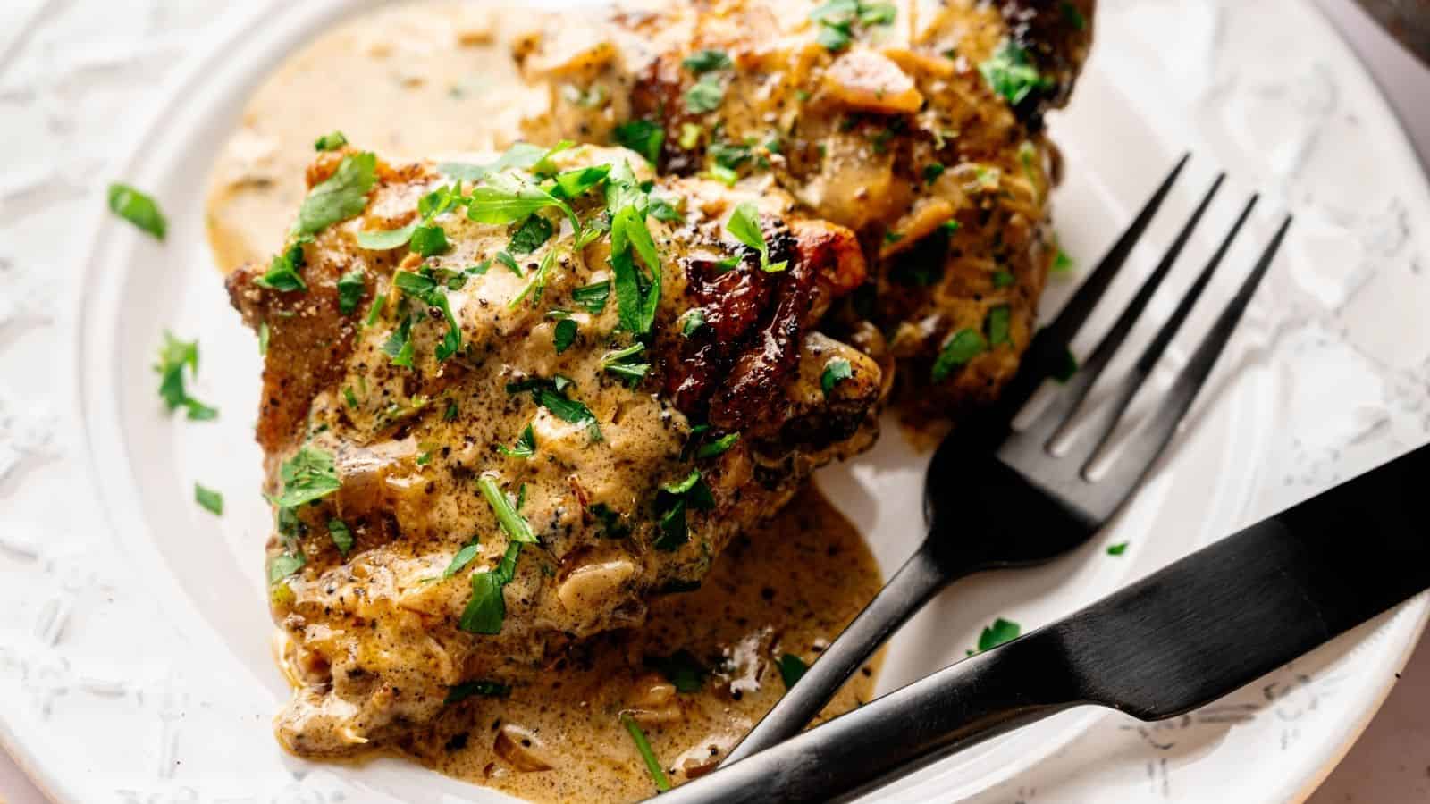 Two pieces of seasoned, creamy chicken garnished with chopped herbs on a white plate, with a black fork and knife beside them.
