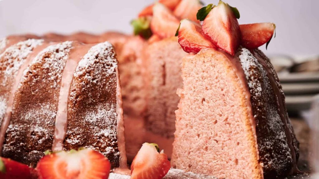 A strawberry Bundt cake topped with sliced strawberries and dusted with powdered sugar, with one slice partially removed.