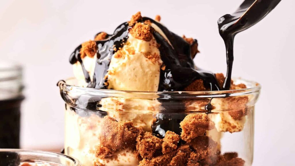 A glass cup filled with vanilla ice cream, crumbled cookies, and chocolate sauce being poured on top.