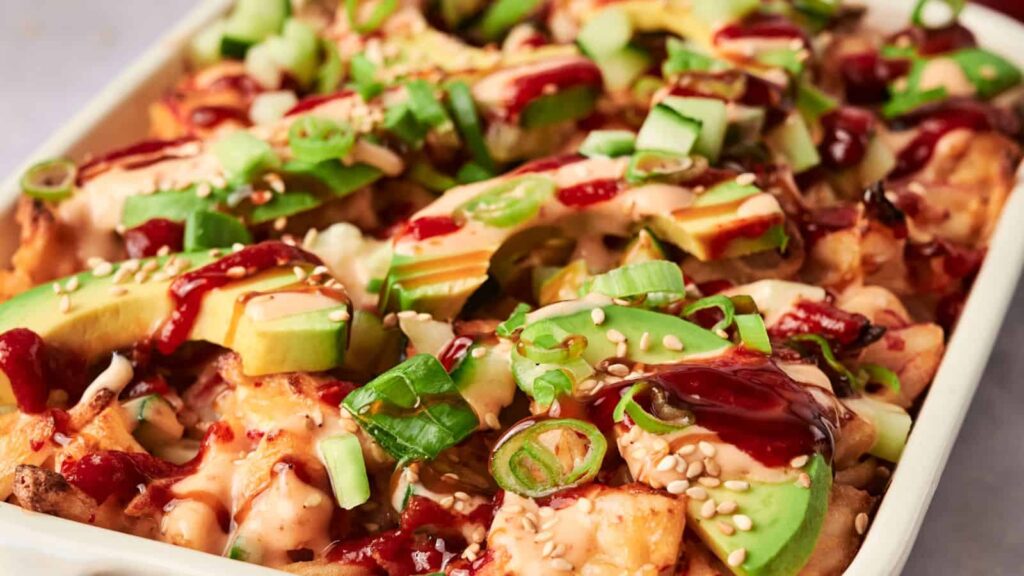 Close-up of a dish topped with sliced avocado, green onions, cucumber, sesame seeds, and drizzled with sauces.