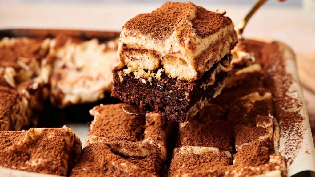 A close-up of a tray of tiramisu with a chocolate base, with one slice being lifted out, topped with dusted cocoa powder.