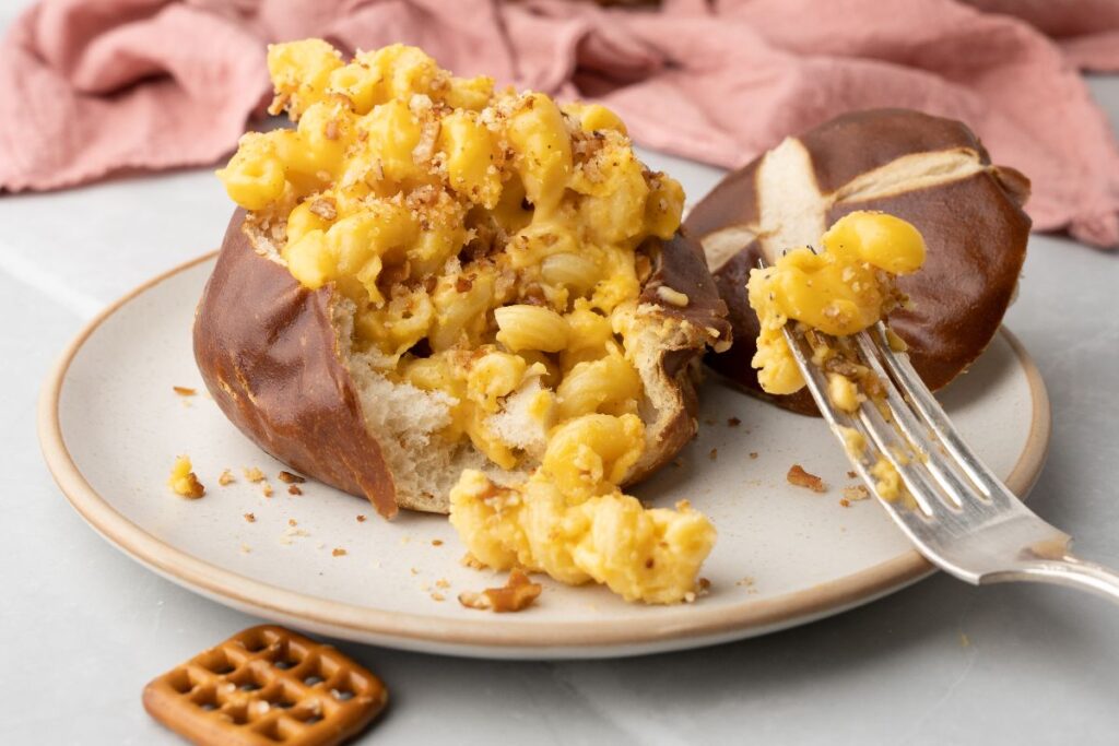 Mac and cheese in a pretzel bun, topped with crunchy panko, served with a fork and pretzel pieces on the plate.