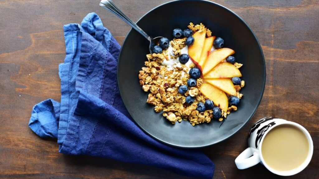 A bowl of maple hazelnut granola with blueberries and peaches in a black bowl on a wooden surface. 