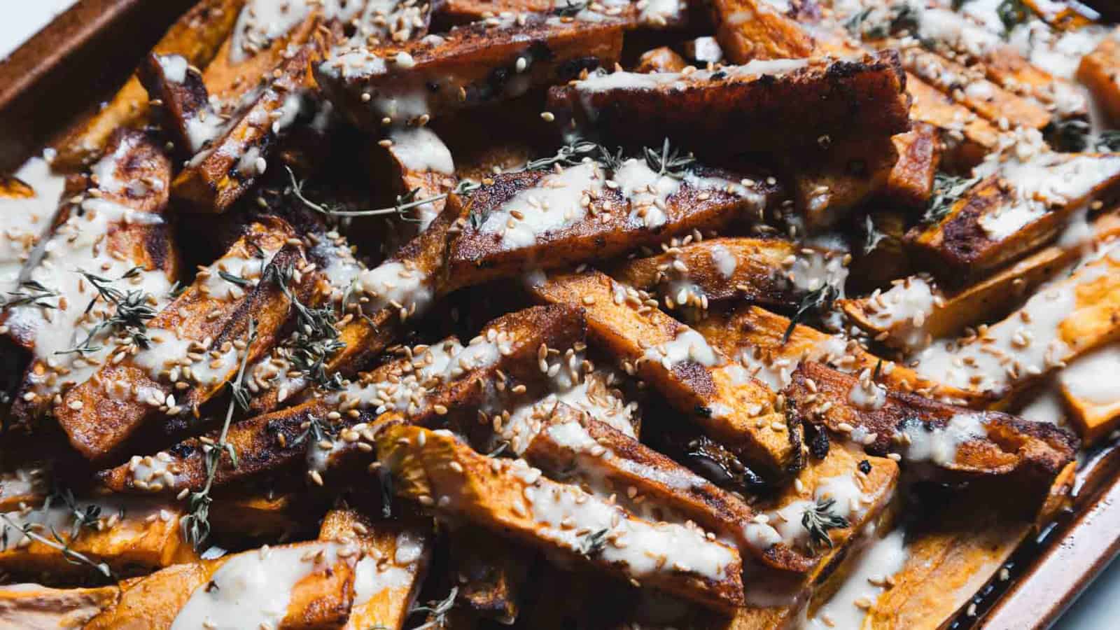 Sweet potato fries topped with a creamy white sauce, herbs, and sesame seeds on a baking tray.