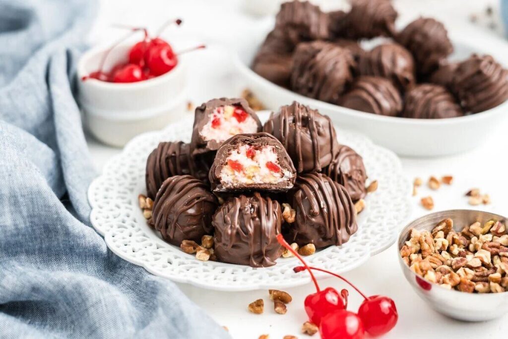 Chocolate truffles with cherry filling sit on a white plate, surrounded by cherries and chopped nuts.