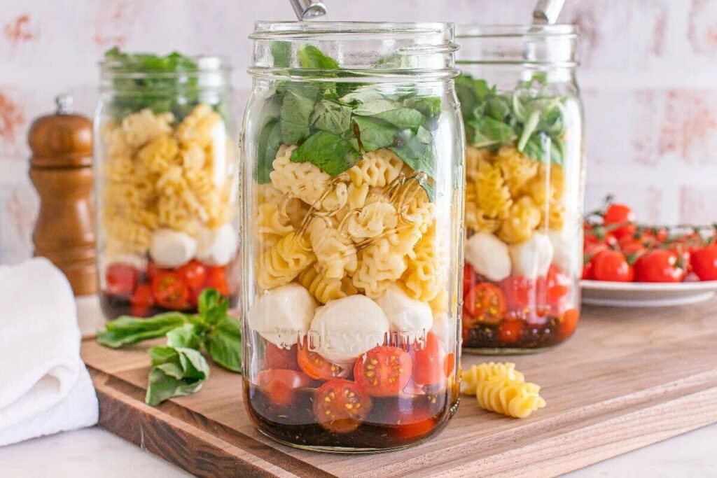 Three mason jars hold Caprese pasta salad with pasta, cherry tomatoes, mozzarella, and fresh basil on a wooden board.