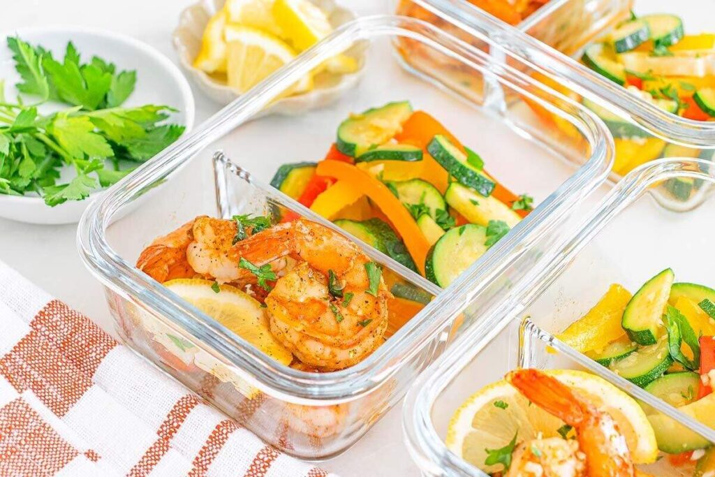 Glass meal prep containers with shrimp, sliced lemon, and sautéed colorful vegetables. 