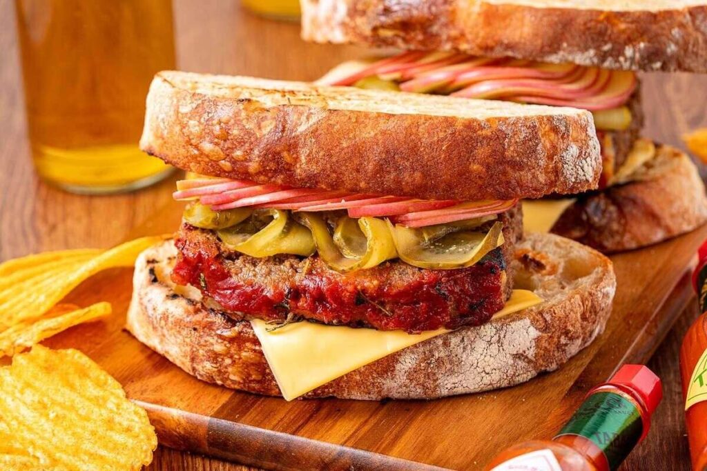 A hearty sandwich with thick bread, melted cheese, meatloaf, pickles, and apple slices on a wooden board.