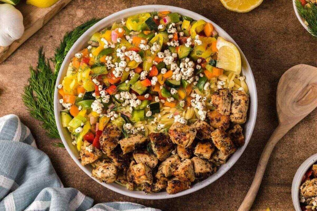 Mediterranean chicken orzo bowls with grilled chicken, vegetables, and grains, topped with herbs and lemon wedges, served with a wooden spoon.