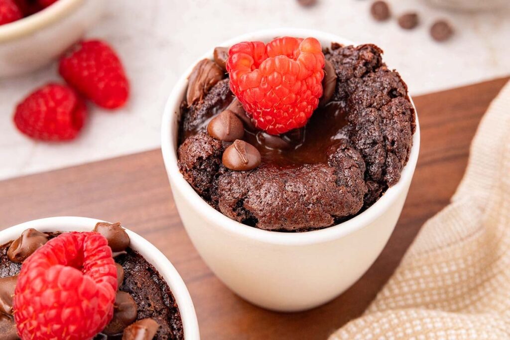 A chocolate mug cake topped with melted chocolate, chocolate chips, and a raspberry, surrounded by extra raspberries and scattered chocolate chips.
