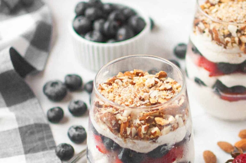 A glass filled with layers of yogurt, blueberries, strawberries, and topped with chopped nuts sits on a white surface with scattered blueberries and a small bowl of blueberries nearby.