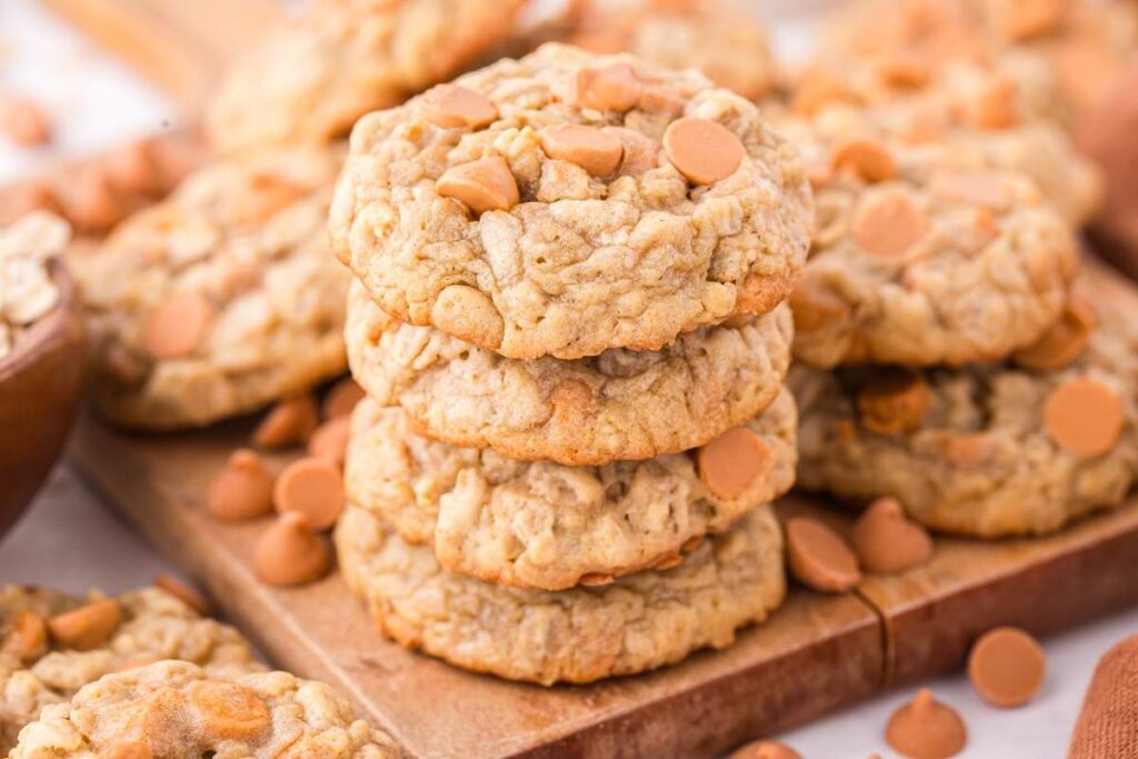 A stack of five oatmeal butterscotch cookies on a wooden surface, with more cookies scattered around.