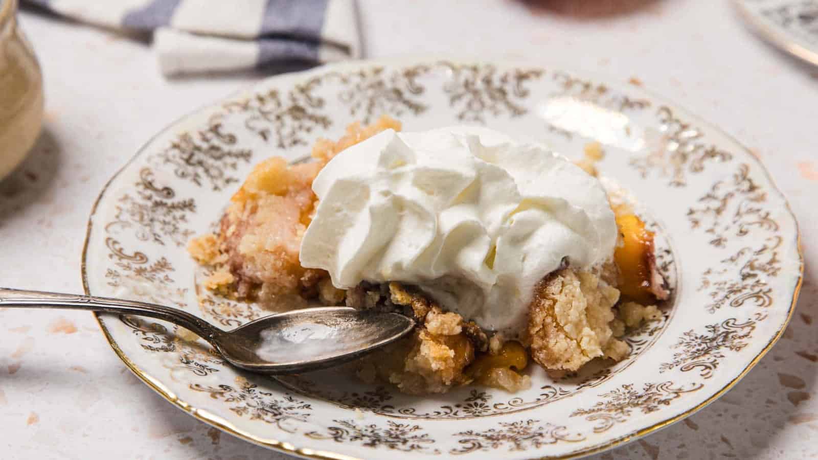 A serving of peach cobbler topped with whipped cream on a decorative plate with a spoon.