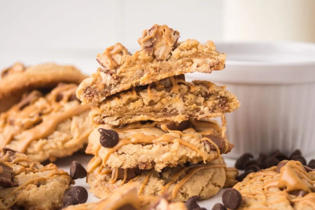 A stack of Peanut Butter Snickers Cookies drizzled with peanut butter and surrounded by chocolate chips.