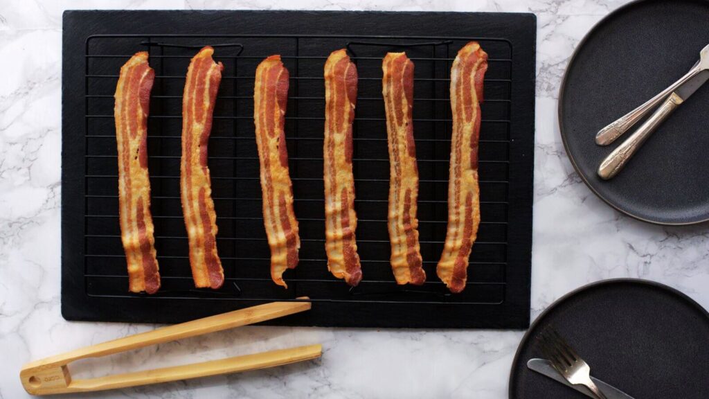 Six strips of cooked bacon rest enticingly on a grill rack, accompanied by wooden tongs and two empty black plates on a marble surface—ready to inspire your next homemade brunch recipe.