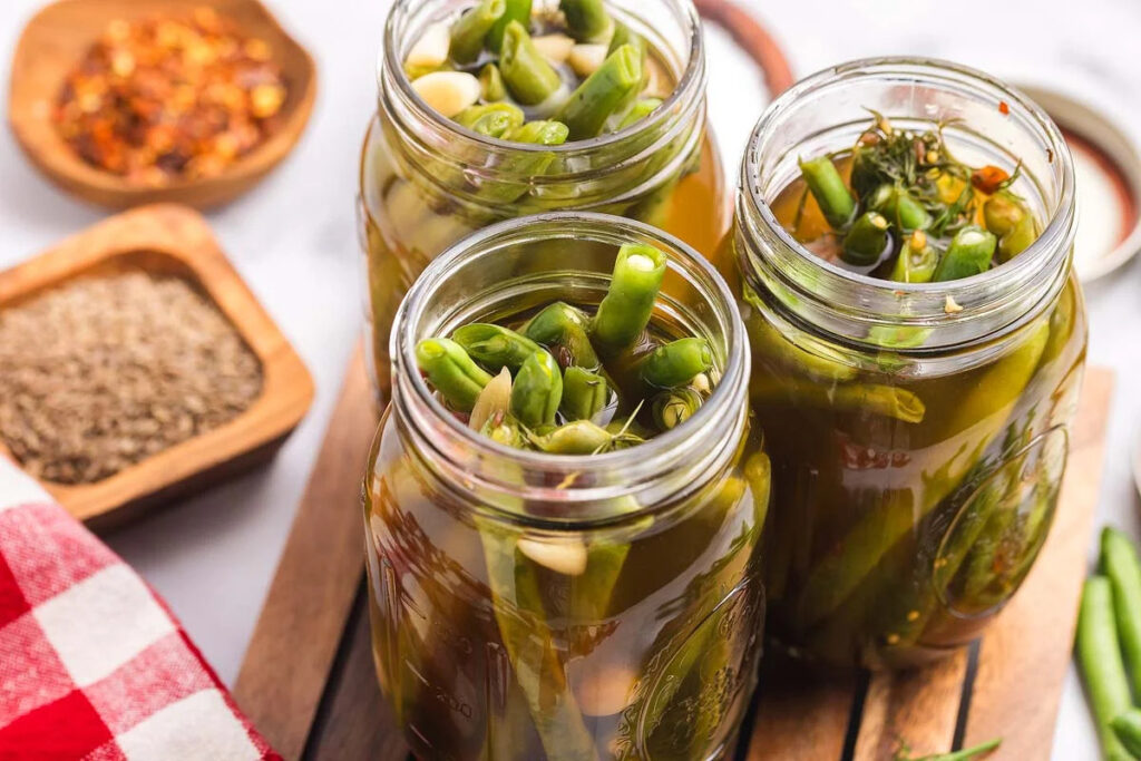 Three jars filled with pickled green beans and garlic sit on a wooden surface.