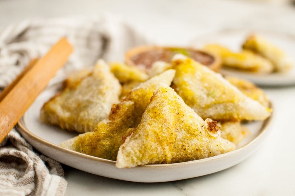 Golden triangular Indian samosas on a white plate with a small bowl of sauce and a striped cloth in the background.