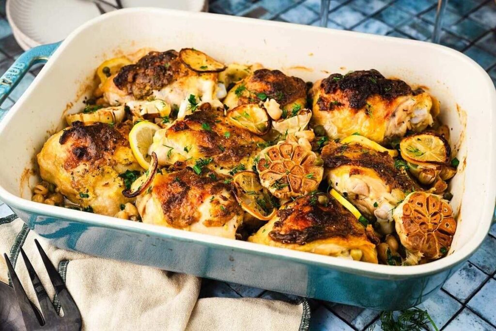 Baked chicken thighs with crispy skin, lemon, roasted garlic, and herbs, served in a blue dish with plates nearby.