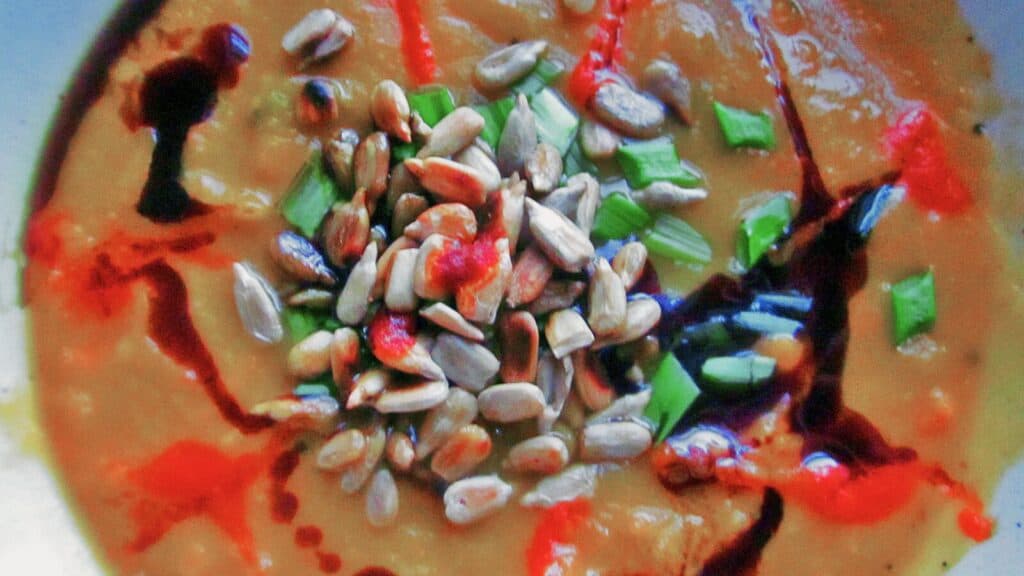 A bowl of soup garnished with seeds, chopped herbs, and drizzles of sauce.