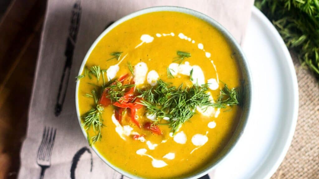A bowl of creamy, hearty orange soup garnished with fresh dill and small pieces of red pepper, drizzled with cream.