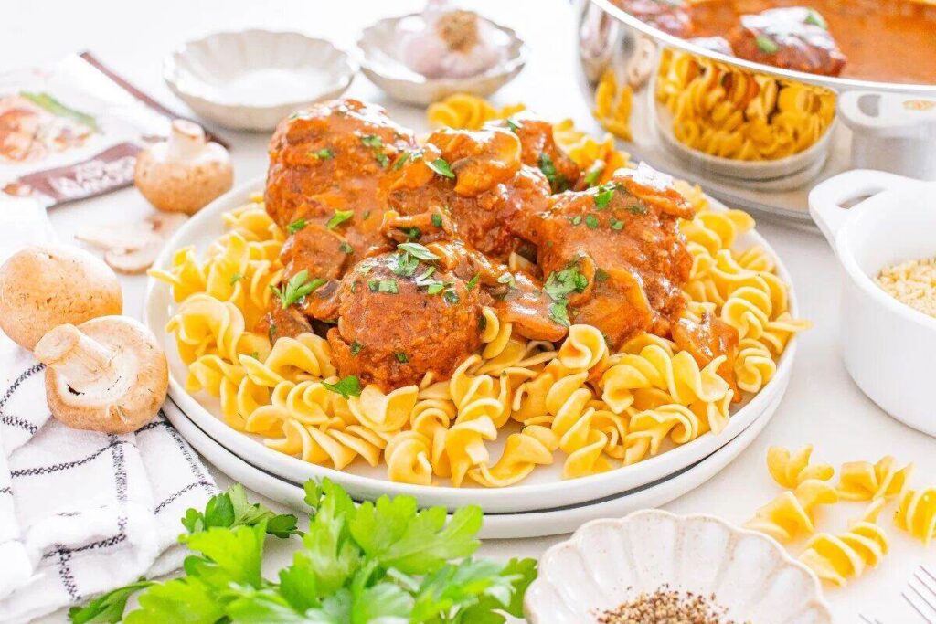 Egg noodles topped with Salisbury steak meatballs in tomato sauce, garnished with herbs and mushrooms.