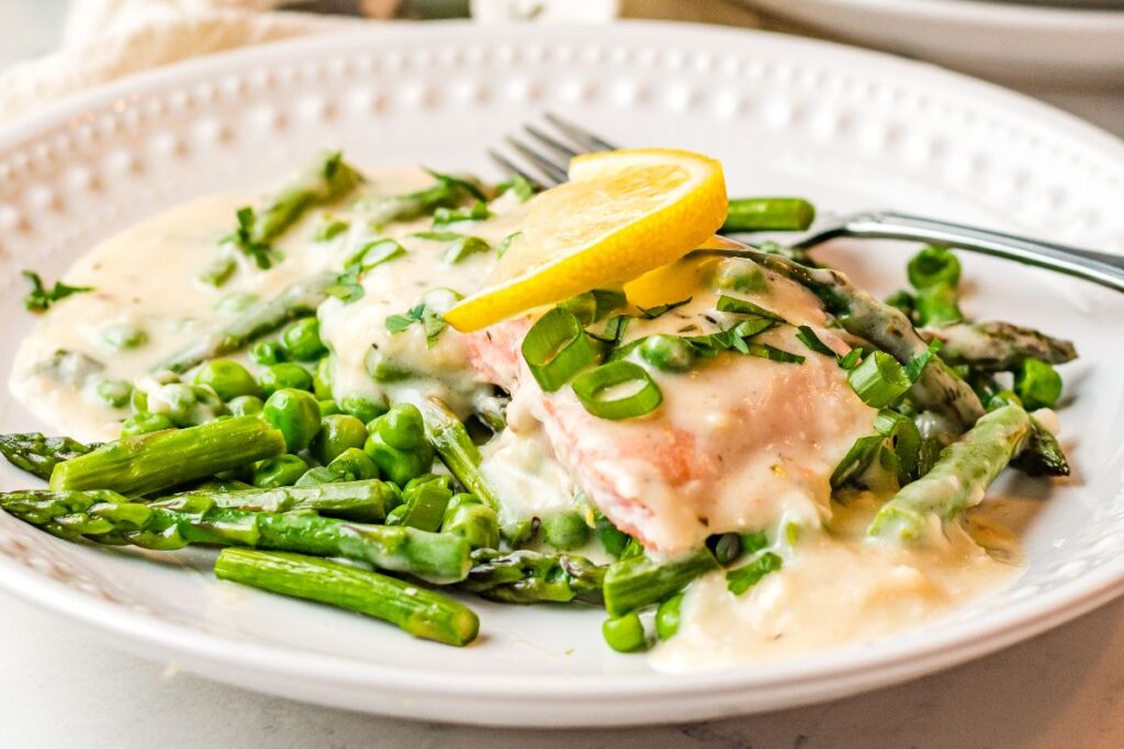 Salmon with white wine sauce, asparagus, and peas, garnished with lemon and herbs, is a simple and elegant dinner idea.