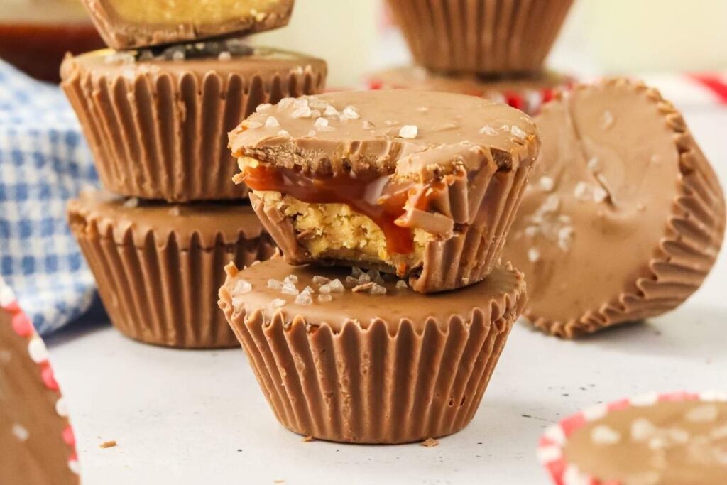 A stack of salted caramel peanut butter cups, with the front one partially bitten to reveal a gooey caramel filling. A sprinkle of sea salt is visible on top of the candies.
