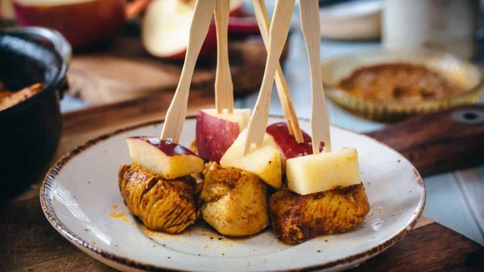 A plate contains skewers with cooked meat cubes and red apple pieces. The skewers are upright, resting on the plate. In the background, there are additional food items and a blurred bowl with a brown dip.