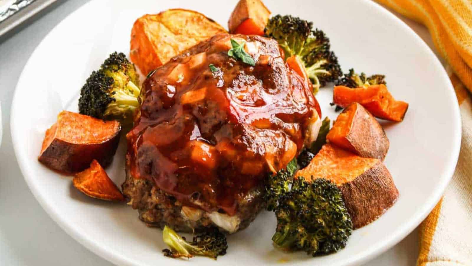 A baking sheet lined with foil holds three glazed meatloaf portions, surrounded by roasted sweet potato cubes and broccoli florets. A yellow and white towel is partially visible to the left.